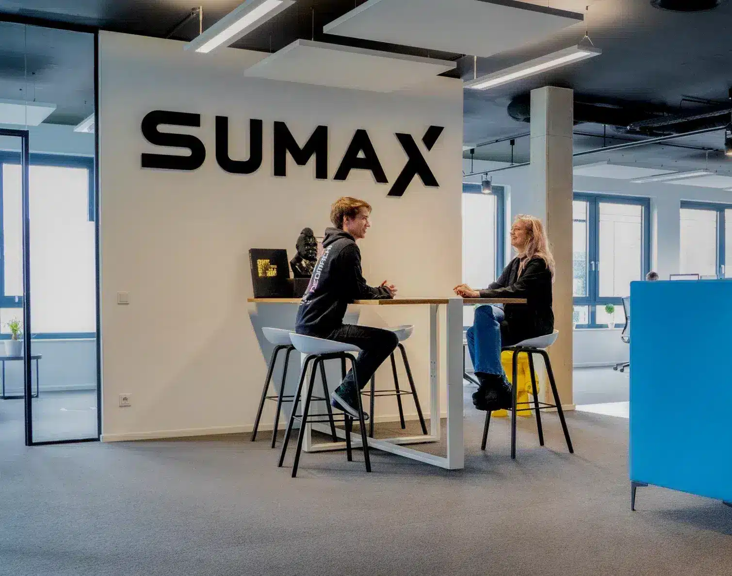 Eingangsbereich des SUMAX-Büros mit Logo an der Wand, zwei Mitarbeitende am Stehtisch